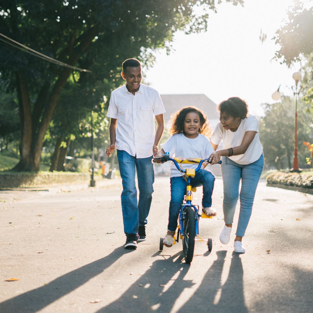 Parents guide their child as she learns to ride a bicycle, encouraging learning new skills to build confidence.