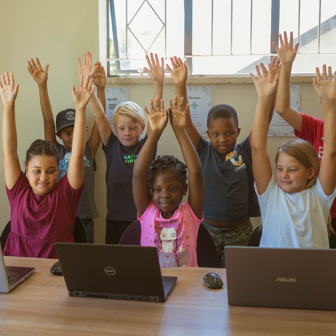 Enthusiastic children raise their arms while learning life skills in a classroom using iRainbow software on their laptops.