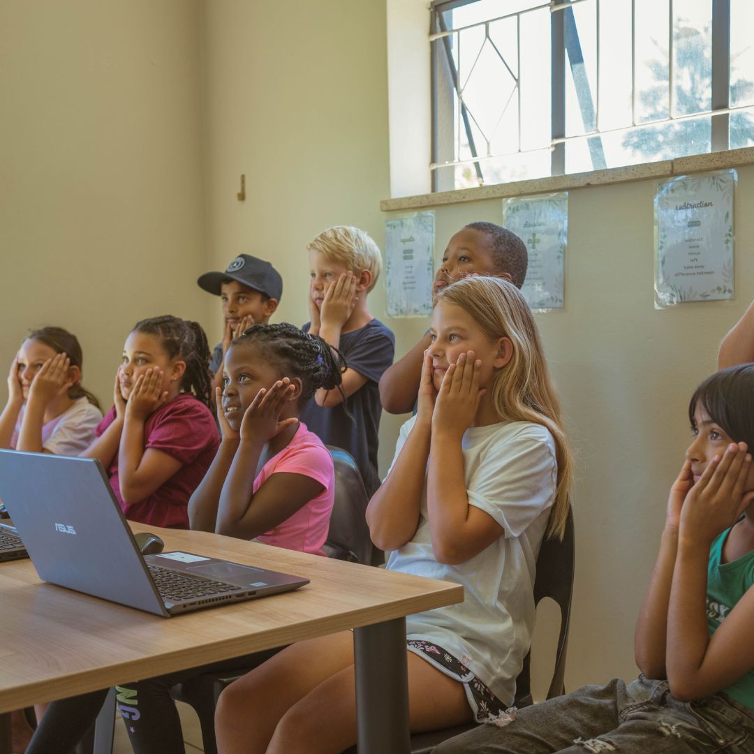 Children are in a classroom with their hands on their faces, learning Grade 1 life skills through iRainbow on laptops.