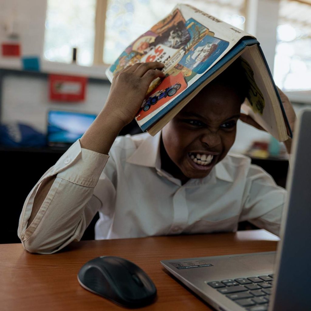 A child is frustrated and stressed looking at a laptop, holding a book on his head