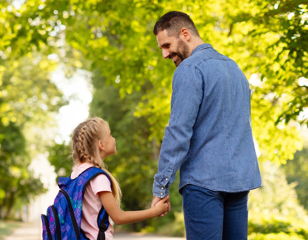Happy middle aged father and daughter schoolgirl walking to school, holding hands and looking at each other