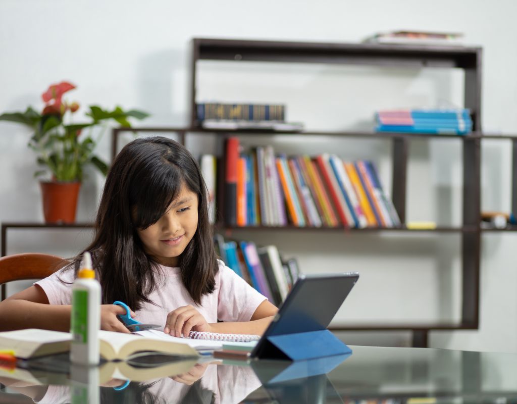 A child is looking at a screen, working with her online homework club. She is holding a scissors and cutting paper.
